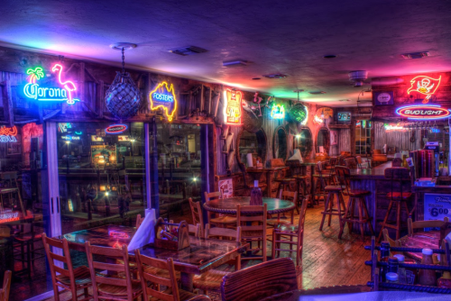 A dimly lit bar with neon signs, wooden tables, and chairs, creating a cozy, inviting atmosphere.
