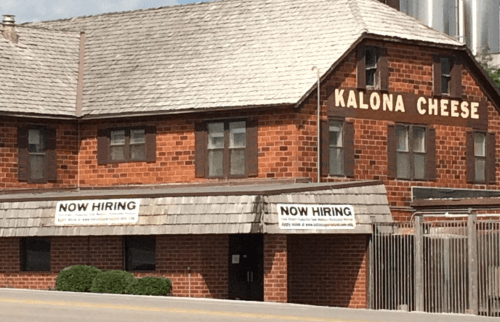 A brick building with a sign reading "Kalona Cheese" and banners stating "Now Hiring" displayed outside.