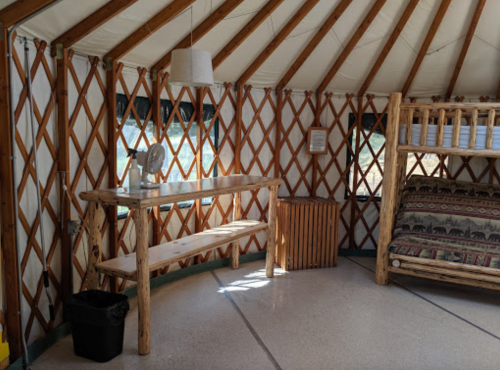 Interior of a yurt featuring wooden furniture, a fan, and a cozy bed with patterned bedding. Natural light filters in.