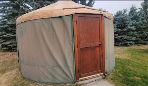 A wooden door on a round, tan yurt surrounded by green grass and evergreen trees.