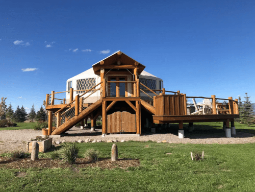 A wooden yurt with a staircase, surrounded by green grass and blue sky, set in a scenic outdoor area.