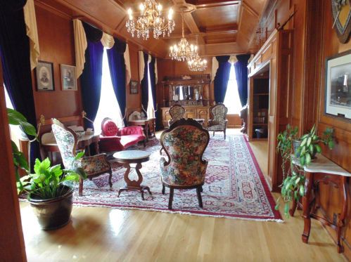 Elegant room with wooden walls, chandeliers, floral chairs, a patterned rug, and large windows with blue curtains.