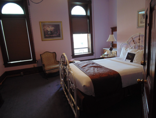 Cozy bedroom with a white metal bed, vintage armchair, and large windows, featuring soft purple walls and wooden accents.
