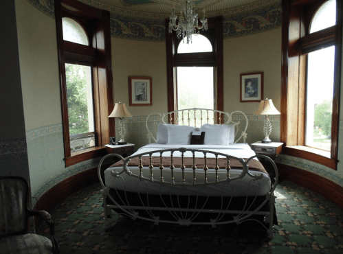 Cozy bedroom with a vintage bed, two lamps, and large windows, featuring floral wallpaper and a patterned carpet.