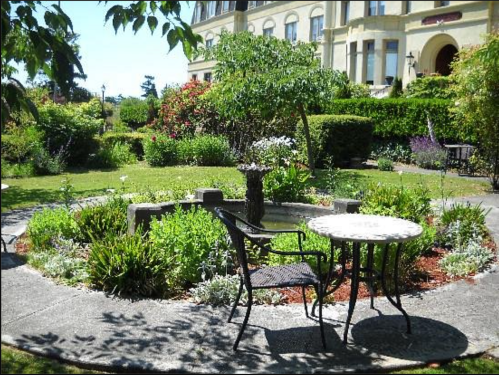 A serene garden with a stone fountain, surrounded by lush greenery and a small table with a chair.