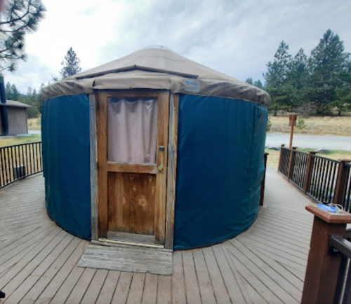 A wooden yurt with a blue exterior, set on a deck surrounded by trees and a grassy area.