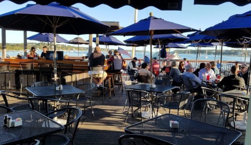 Outdoor dining area with blue umbrellas, tables, and patrons enjoying a waterfront view.