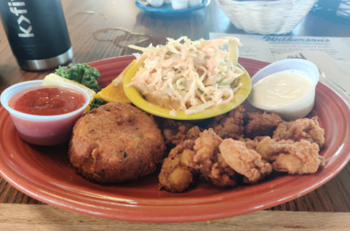 A plate of fried seafood, a crab cake, coleslaw, and dipping sauces, garnished with lemon and kale.