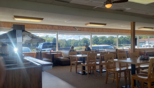 Interior of a restaurant with wooden tables and chairs, large windows, and two people sitting at a booth.