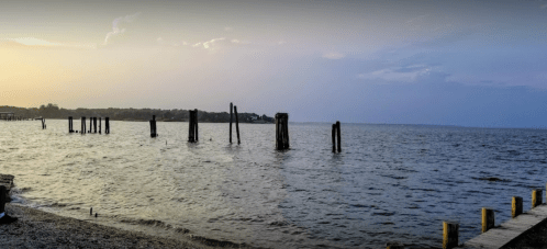 Sunset over a calm lake with weathered wooden posts emerging from the water and a distant shoreline.