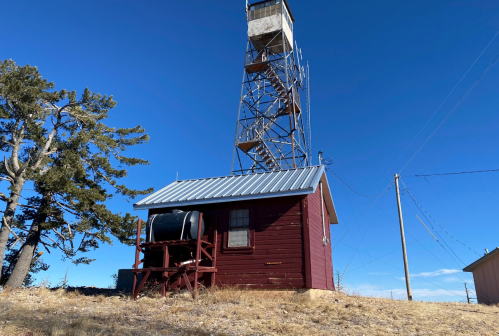 A red wooden building with a metal roof next to a tall fire lookout tower against a clear blue sky.