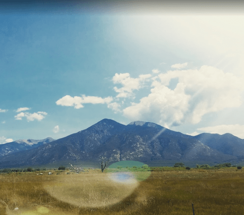 A scenic view of mountains under a blue sky with fluffy clouds, surrounded by a grassy field. Sunlight creates a lens flare effect.