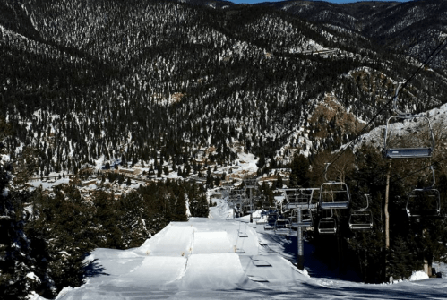 A snowy ski slope with chairlifts, surrounded by tall pine trees and mountains in the background.