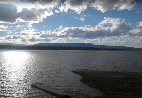 A serene lake scene with a dock, mountains in the background, and a sky filled with clouds reflecting sunlight.