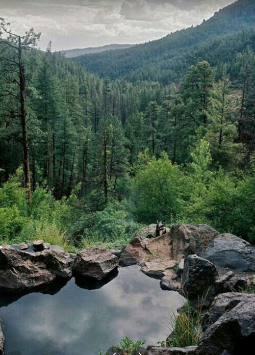 A serene natural landscape featuring a rocky pond surrounded by lush green trees and mountains under a cloudy sky.