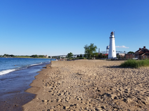 A sandy beach with a lighthouse in the background under a clear blue sky. Waves gently lap at the shore.