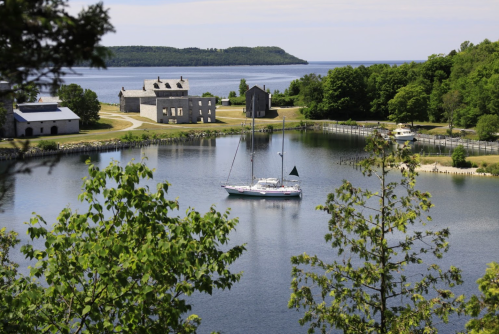 A serene harbor scene with a sailboat anchored near historic buildings and lush greenery under a clear sky.