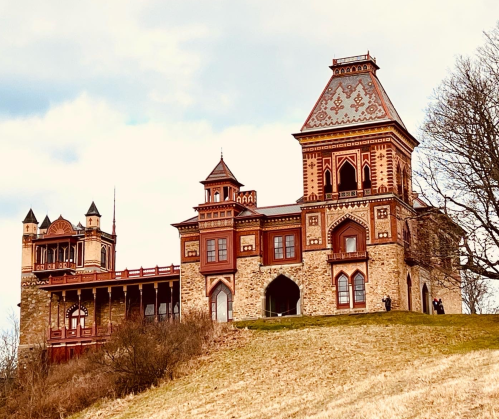 A historic castle with intricate architecture, set on a grassy hill under a cloudy sky.