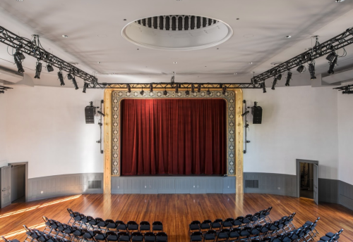 A spacious theater with a red curtain, wooden floor, and rows of black chairs facing the stage.
