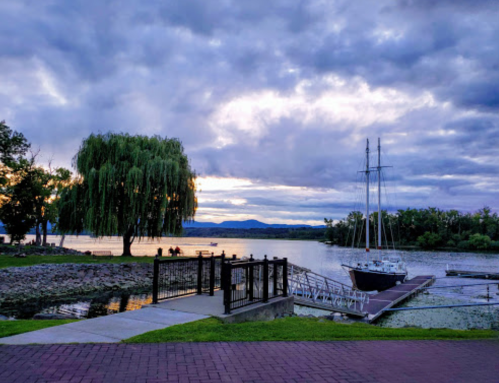 A serene waterfront scene with a docked sailboat, lush trees, and a cloudy sky reflecting on the water.