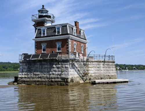 A historic lighthouse on a stone base, surrounded by water, with a clear blue sky in the background.