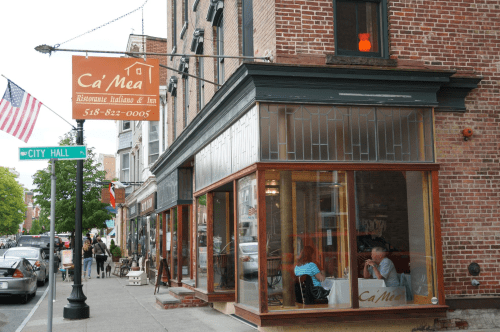 A street view of Ca'Mea restaurant with large windows, showcasing diners inside and a city hall sign nearby.