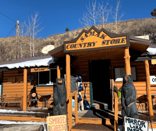 A rustic country store with bear statues at the entrance, surrounded by snow and trees on a sunny day.