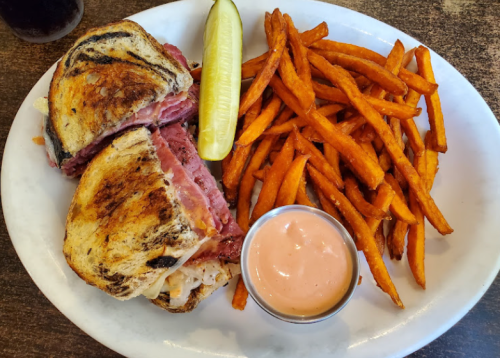 A plate with a toasted sandwich filled with meat, a pickle, and a side of sweet potato fries with dipping sauce.