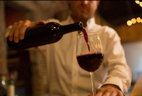 A bartender pours red wine into a glass, with a softly lit background and warm ambiance.