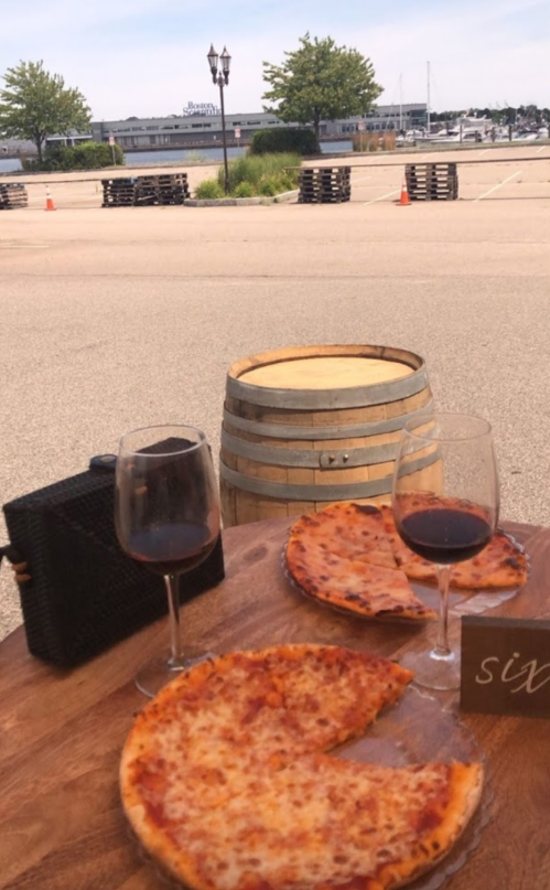 A wooden table with two glasses of red wine and two pizzas, with a scenic view of a harbor in the background.