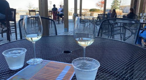 Two glasses of white wine and a cup of water on a table, with people dining in the background on a sunny day.