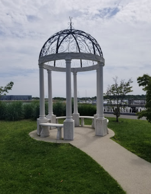 A white gazebo with a decorative dome, surrounded by grass and trees, near a waterfront with boats in the background.