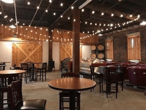 Cozy winery interior with wooden beams, string lights, tables, and wine barrels in the background.
