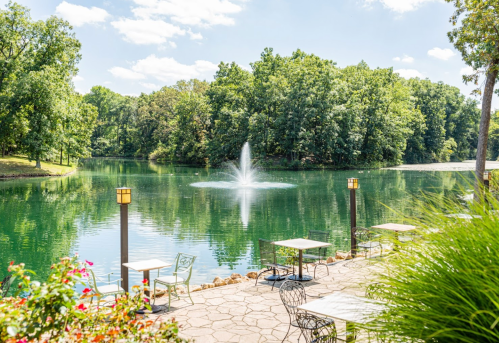 A serene lake scene with a fountain, surrounded by lush greenery and outdoor seating along the water's edge.