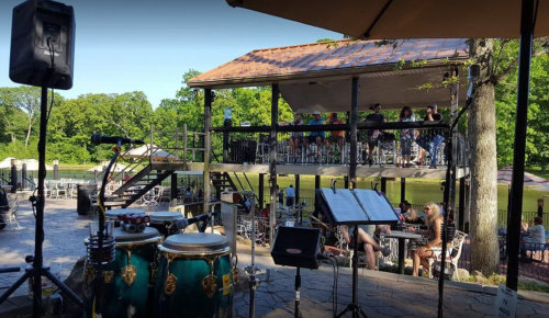 A lively outdoor venue with a stage, seating area, and musicians' instruments in the foreground, surrounded by greenery.