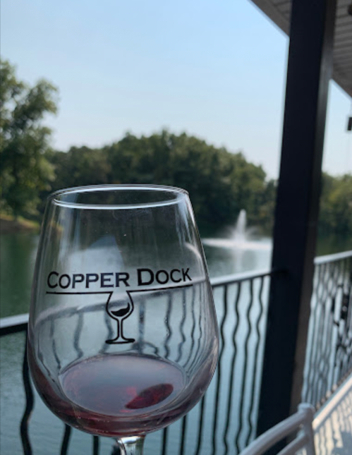 A glass of red wine with "Copper Dock" logo, overlooking a serene lake and trees in the background.