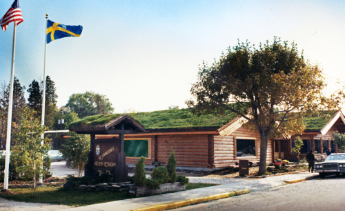 A log cabin-style building with a green roof, featuring flags and a sign, surrounded by trees and a parking area.