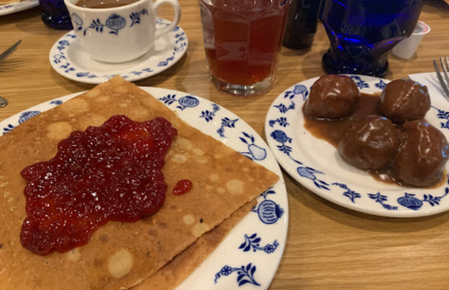 A plate with a large crepe topped with jam, alongside a plate of sweet dumplings in sauce and a cup of coffee.