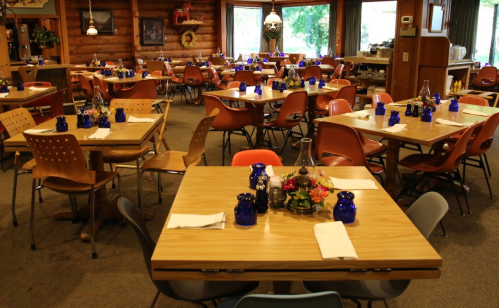 A cozy dining area with wooden decor, tables set with blue glass candle holders, and colorful chairs.