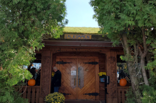 A wooden building entrance with a green roof, featuring a sign that says "Valkommen" and surrounded by greenery and pumpkins.