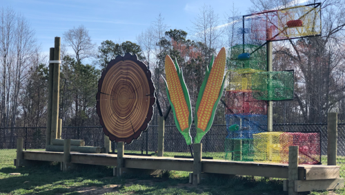 Colorful playground structure featuring a log, corn, and climbing nets against a clear blue sky.