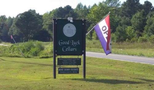 Sign for Good Luck Cellars with an open flag, surrounded by greenery and a road in the background.