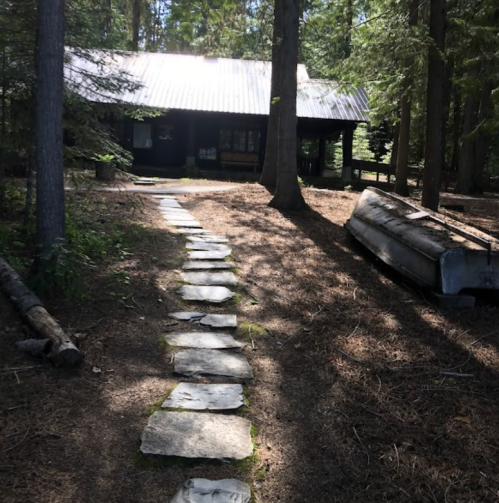 A stone path leads to a dark cabin surrounded by tall trees and a canoe resting on the ground.
