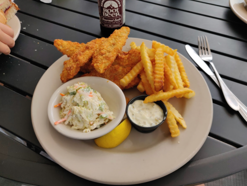 A plate with crispy fried fish fillets, crinkle-cut fries, coleslaw, a lemon wedge, and a bottle of root beer.