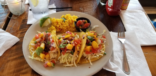 A plate of three tacos topped with fresh salsa, served with yellow rice and a lime wedge on a wooden table.