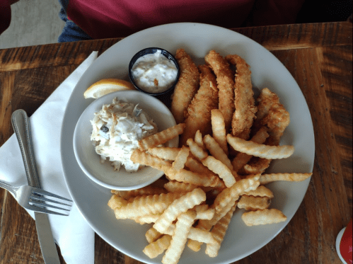 A plate of crispy fried fish fillets, crinkle-cut fries, coleslaw, and a lemon wedge, served with dipping sauce.