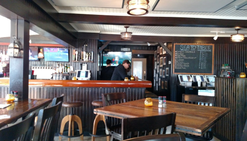 Interior of a rustic restaurant with wooden tables, a bar, and screens showing sports. Staff are serving customers.