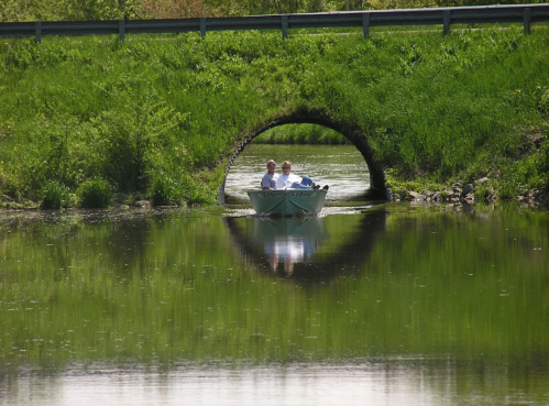 Two people in a small boat pass under a stone arch bridge, surrounded by lush greenery and calm water reflections.