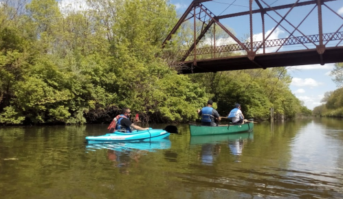 Two people kayak on a calm river beneath a metal bridge, surrounded by lush green trees.