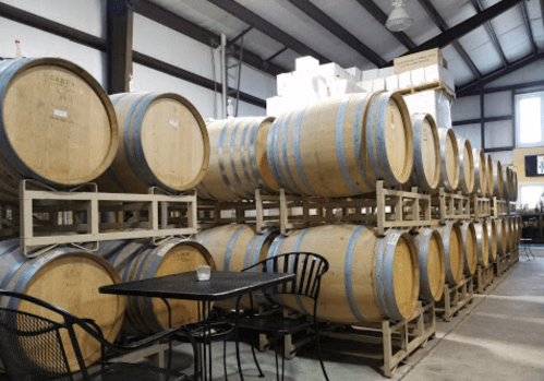 A row of wooden wine barrels stacked in a warehouse, with a small table and chairs in the foreground.